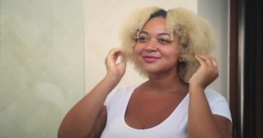 Kısa kıvırcık sarı afro saçlı, genç Afrikalı Amerikalı kadın aynadaki yansımasına bakar ve hayran kalır. Kendini beğenme ve kendini kabullenme. Vücut pozitif. Cilt ve saç bakımı.