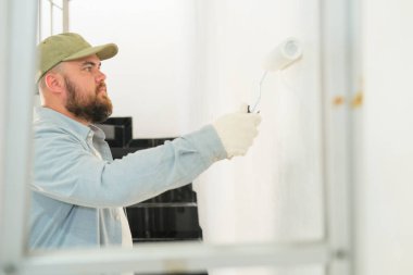 İnşaat eldivenli bir adamın merdiven boşluğundan beyaz boyayla silindir kullanarak duvarı boyadığını görmek. Yeni ev tadilatı, yeni bir eve taşınma hazırlığı. Profesyonel ressam. İşçi.