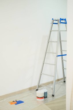 Yeni bir eve taşınmadan önce bir daire tadilatı. Bir merdiven beyaz bir duvara dayanır, yanında boya kutuları, bir spatula, bir silindir ve iş için fırçalar vardır..