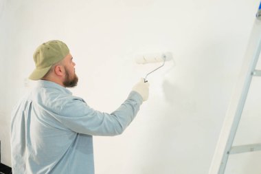 Sakallı ve inşaat eldivenli bir adam silindir kullanarak duvarı beyaz boyayla boyuyor. Yanında bir merdiven ve diğer aletler var. Yeni bir evde tamirat, yeni ev partisi hazırlıkları..