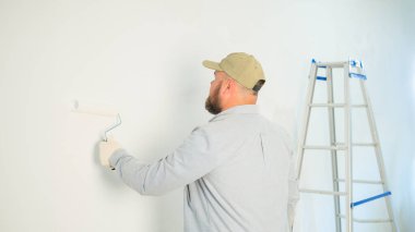 İnşaat eldivenli bir adamın beyaz boyalı bir duvarı arka planda merdiven kullanarak boyadığını gördüm. Yeni bir evin yenilenmesi, yeni bir eve taşınma hazırlığı. Profesyonel..