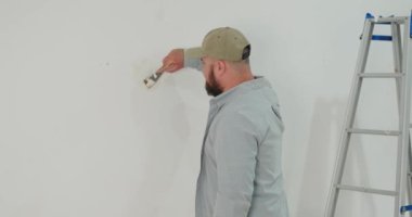 Sıradan kıyafetlerle duvardan eski boyaları spatulayla kazıyan bir adamın yan görüntüsü. Arka planda bir merdiven duruyor. Duvarları boyamaya hazırlıyorum. Yeni bir evde yenileme işi.