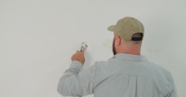 Tadilata başlamadan önce elinde spatula tutan ve duvardan beyaz boya kazıyan şapkalı bir adamın arka planı. Duvarları boyamaya hazırlıyorum. Bir apartmanın içinde tadilat işi.