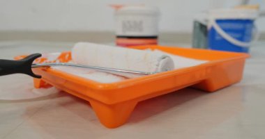 Arka planda beyaz boyalı plastik bir tepside duran silindirli bir arka plan videosu var. Arka planda boya kutuları var. Yeni bir evin içinde onarım. Duvarları boyama süreci. İnsanlar olmadan.