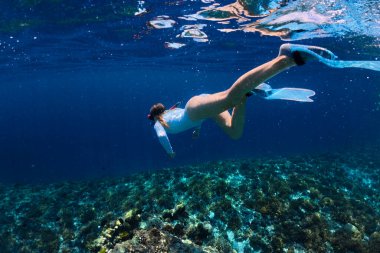 Mavi dalgıç kıyafetli, yüzgeçli ve maskeli genç bir kadın suyun altında yüzüyor ve tropik adanın yakınlarındaki mercan resiflerine ve balıklara bakıyor. Tatil sırasında şnorkelle yüzme ve serbest dalış aktiviteleri.