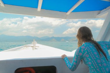 Ahşap bir teknenin güvertesinde oturmuş dağları olan tropik bir adaya bakan bir kadının arka görüntüsü. Tekne okyanustaki dalgalar boyunca hareket ediyor. Adaya atlama ve dalış turu