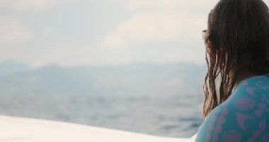 Ahşap bir teknenin güvertesinde oturmuş dağları olan tropik bir adaya bakan bir kadının arka görüntüsü. Tekne okyanustaki dalgalar boyunca hareket ediyor. Adaya atlama ve dalış turu