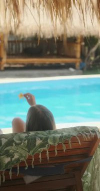 Mayo giymiş genç sarışın bir kadın havuzun yanındaki güneş pansiyonunda uzanıyor ve elinde plumeria çiçeği tutuyor. Bungalovdaki tropik cennette tatil, çiçekleri koklayan, gülümseyen ve suda dinlenen bir kadın.