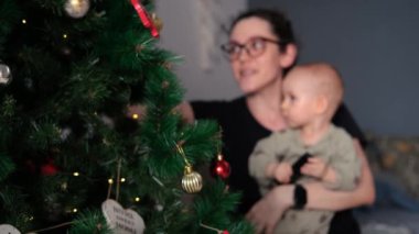 Ön planda süslü bir Noel ağacı var. Arka planda, bir anne ve küçük kızı bir kanepede oturuyor, ağaçtaki süslemelere bakıyor ve gülümsüyorlar. İyi tatiller..