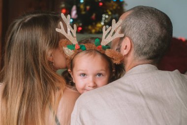 Noel ağacının arka planında, anne, baba ve küçük kızları sarılıyorlar, boynuzları boynuzlu saç bandı takıyorlar. Kız gülüyor ve mutlu bir şekilde ailesine sarılıyor. Aile tatili.