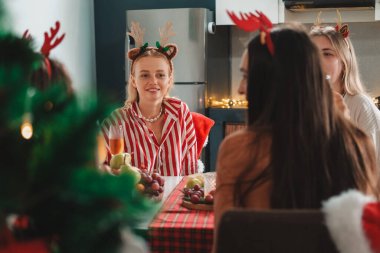 Geyik boynuzlu saç bandı takan genç bayan arkadaşlar mutfak masasında oturmuş Noel 'i kutluyorlar. Şampanya bardaklarını tokuşturup gülümserler. Yakınlarda bir Noel ağacı var..
