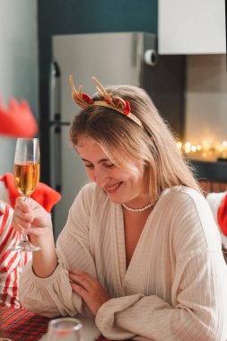 Mutfak masasında oturan genç sarışın bir kadının portresi geyik boynuzlu bir saç bandı takıyor ve Noel 'i yakın arkadaşlarıyla kutluyor. Şampanya içip gülümsüyor..