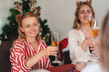 Geyik boynuzlu saç bandı takan genç bayan arkadaşlar mutfak masasında oturmuş Noel 'i kutluyorlar. Şampanya bardaklarını tokuşturup gülümserler. Yakınlarda bir Noel ağacı var..