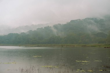 Gün batımında dağlarda bir göl manzarası, her yer sis. Mistik bir atmosfer. Tahta balıkçı tekneleri yakınlarda, ama hiç insan yok. Suyun yakınındaki ağaçlar ve ormanlar.