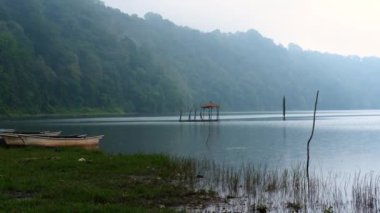 Gün batımında dağlarda bir göl manzarası, her yer sis. Mistik bir atmosfer. Tahta balıkçı tekneleri yakınlarda, ama hiç insan yok. Suyun yakınındaki ağaçlar ve ormanlar.