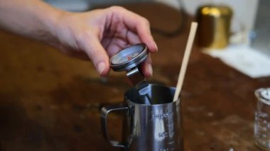 Kadın ellerinin, sobadaki demir kasede erimiş mum ısısını ölçen yakın plan. Atölyede el yapımı kokulu mum yapma süreci.