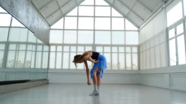 Geniş ve parlak bir stüdyoda, bir kadının zarif, çağdaş bir dans gösterisi sergilediği geniş açılı bir çekim. Hareketleri zarafet ve esnekliği birleştirir..