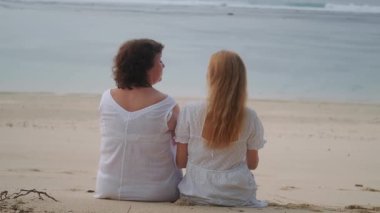 Bir aile tatili sırasında sahilde oturan yaşlı bir anne ve genç kızıl saçlı kızının arka görüntüsü. Sarılırlar ve okyanusa bakarlar. Anneler Günü.