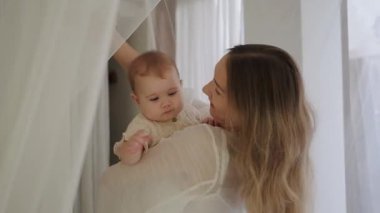 Küçük kızını kucağında tutan ve onunla oynayan genç bir kadın. Sevimli bebek mutlu bir şekilde gülümsüyor. Bebeği uyutmak için sallıyorum. Anneler Günü. Aşk ve sevgi.