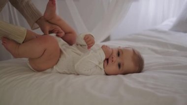 Bir bebek beyaz bir battaniyenin üzerinde yatağa uzanır, annesinin elleri çocuğu giydirir ve elbiselerini bağlar. Küçük bir kız yatakta gülüyor ve dönüyor. Doğum izni mi? Anneler Günü. Sevgi ve ilgi.