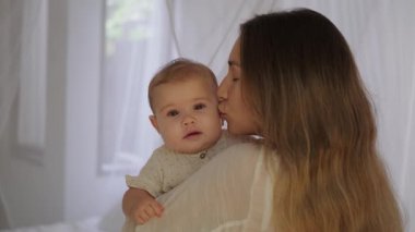 Genç bir kadın küçük kızını kollarından tutar ve şefkatle öper. Sevimli bebek mutlu bir şekilde gülümser. Bebeği uyutmak için sallıyor. Anneler Günü. Aşk ve sevgi.
