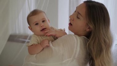 Küçük kızını kucağında tutan ve onunla oynayan genç bir kadın. Sevimli bebek mutlu bir şekilde gülümsüyor. Bebeği uyutmak için sallıyorum. Anneler Günü. Aşk ve sevgi.