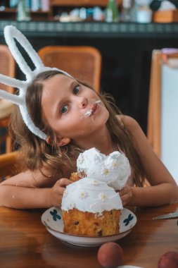 Kafasında tavşan maskesi olan bir kız paskalya pastasını mutfak masasında yiyor. Pastadan bir parça ısırıyor ve beyaz krem burnunda ve dudaklarında kalıyor. Tatlı bayram tatlısı. Aile gelenekleri.
