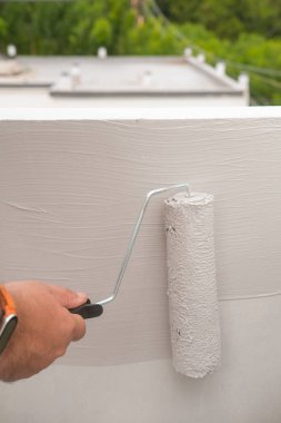 Patenci tutan ve çatıya gri boyalı bir duvar çizen bir adamın yakın çekimi. Tamircinin biri yazın ikinci kattaki evin dış cephesini yeşilliğin arka planına boyuyor.
