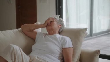 Beyaz saçlı son sınıf öğrencisi bir kadın oturma odasındaki kanepede oturuyor ve kafasını ellerinin arasında tutuyor. Başı ağrıyor ve iyi hissetmiyor. Su içer ve eliyle yüzünü kapatır. Tıbbi bakım.