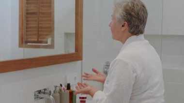 Son sınıf öğrencisi bir kadın banyoda aynanın önünde duruyor ve yüzüne masaj hareketleriyle nemlendirici kırışıklık önleyici krem sürüyor. Yaşlanan cilde dikkat et. Emeklilik yaşında güzellik rutini.