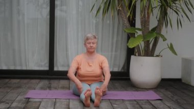 Eşofmanlı gri saçlı son sınıf öğrencisi bir kadın tropikal bir villada yüzme havuzlu bir yerde yoga yapıyor. Yaşlılar için sağlık jimnastiği. Emeklilikte dinlenme ve iyileşme.