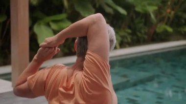 Yüzme havuzunun yanındaki tropikal bir villada elleri için terapi egzersizi yapan gri saçlı yaşlı bir kadının arkası. Emeklilikte dinlenme ve iyileşme.