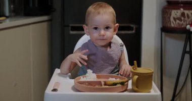 Küçük bir kız yüksek bir sandalyeye oturur ve öğle yemeğini yer. Bebek çatalla yemek yemeyi öğreniyor, ama pilavı eliyle alıp ağzına koyuyor. Yiyeceklerle oynuyor ve eğleniyor..