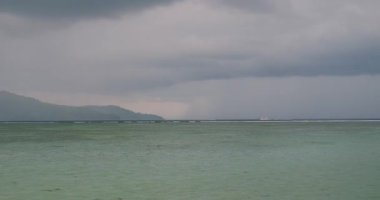 Adadaki kumsaldan okyanus manzarası, mavi su ve küçük dalgalar, bulutlu hava, dağlar uzaktan görülebilir. Hızlı tekneler uzaklarda hareket ediyor, turistleri taşıyor..
