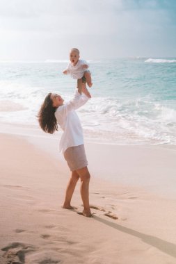 Plajda kucağında bir bebekle yürüyen genç bir kadının portresi. Bebeği arka planda okyanus varken havaya fırlatıyor ve bebek şaşırıyor. Ebeveynlerle ilk çocuk tatili. 
