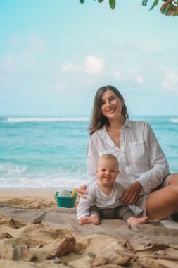 Kumsalda oturup oyuncaklarla, kürekle ve kovayla oynayan bir bebeğin portresi. Sahilde tatil yapan çocuklar için eğlence. Yanında annesi oturuyor ve ona yardım ediyor. Anneler Günü.