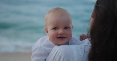 Annesi tarafından okyanusa karşı tutulan mavi gözlü bir bebeğin yakın plan portresi. Anne oğlunun yanağından öpüyor, çocuk gülüyor. Çocuklu bir tatil. Anneler Günü. Aile seyahati.
