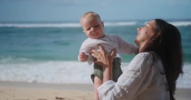 Okyanus kıyısındaki sahilde tatil yapan anne ve bebeğin portresi. Küçük çocuk kaprisli ve ağlıyor, anne onu sakinleştiriyor öpüyor ve neşelendirmek için gıdıklıyor. Anneler Günü. Çocukla seyahat et.