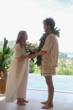 Arka planda yeşil tepeler ve okyanus ile açık hava düğün töreni. Beyaz elbiseli gelin ve damat, çiçek kemerinin ortasında el ele tutuşuyorlar. Evlilik yemini etmek. Aşk.