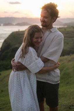 Genç kadın ve erkek doğada gün batımına karşı sarılıyorlar. Balayı gezisi, düğünden sonra aşık bir çift. Tepedeki okyanusta romantik bir randevu. Sevgililer Günü. Aşk ve romantik ilişkiler.