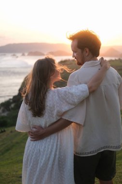 Genç kadın ve erkek doğada gün batımına karşı sarılıyorlar. Balayı gezisi, düğünden sonra aşık bir çift. Tepedeki okyanusta romantik bir randevu. Sevgililer Günü. Aşk ve romantik ilişkiler.