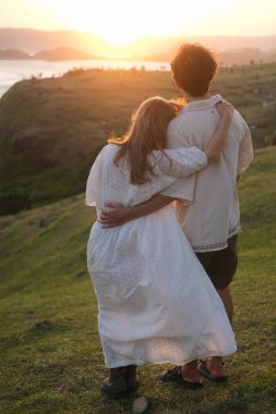Genç kadın ve erkek doğada gün batımına karşı sarılıyorlar. Balayı gezisi, düğünden sonra aşık bir çift. Tepedeki okyanusta romantik bir randevu. Sevgililer Günü. Aşk ve romantik ilişkiler.