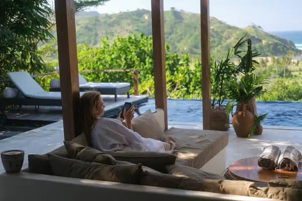 Bornozlu genç bir kadın sonsuz havuzlu lüks bir villanın terasında oturuyor ve elinde bir telefon tutuyor. Sosyal medya yayınlarını inceliyor. Tropik bir tatilde bir villada tatil.