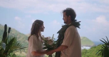 Arka planda yeşil tepeler ve okyanus ile açık hava düğün töreni. Beyaz elbiseli gelin ve damat, çiçek kemerinin ortasında el ele tutuşuyorlar. Evlilik yemini etmek. Aşk.