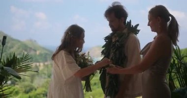 Arka planda yeşil tepeler ve okyanus ile açık hava düğün töreni. Beyaz elbiseli gelin ve damat, çiçek kemerinin ortasında el ele tutuşuyorlar. Evlilik yemini etmek. Aşk.