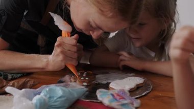 Bir kadın pasta şefi çocuklara Noel kurabiyesini nasıl süslemeleri gerektiğini öğretiyor. Bir pasta poşeti tutuyor ve çocuklara göstererek zencefilli ekmeğin üzerine resim çiziyor. Çocuklar için şenlik atölyesi