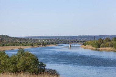 Dniester. Demiryolu köprüsünden nehrin manzarası. Mavi gökyüzü suya yansıyor. Yeşil ağaçlar ve kuru sazlıklar. Yatay fotoğraf.