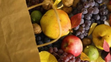 Elma, armut, ceviz, kuru erik ve üzümlerin sonbahar hasadı bir kese kağıdından masaya dökülür, yanında da canlı düşen yapraklar. Odak değişimi, zengin sonbahar renkleriyle birlikte doğal ve çevre dostu ambalajı vurguluyor.