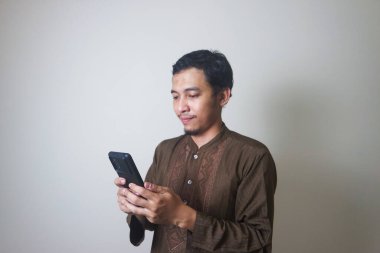 Neşeli Asyalı genç Müslüman bir adam cep telefonu kullanıyor ve beyaz arka planda izole bir şekilde yüzünü çeviriyor.
