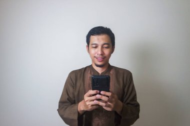 Neşeli Asyalı genç Müslüman bir adam cep telefonu kullanıyor ve beyaz arka planda izole bir şekilde yüzünü çeviriyor.